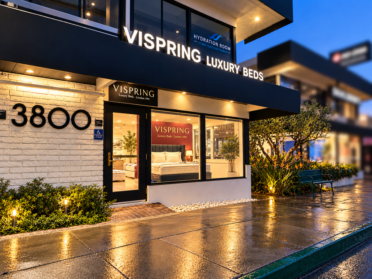 Front exterior of a Vispring Luxury Beds retail store at dusk, with illuminated signage, glass windows, wet pavement reflecting warm lights, and landscaped shrubs along the sidewalk.